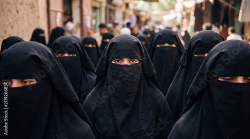 A group of Afghan women wearing traditional black burkas and face veils walking together in a crowded market street, representing Islamic culture, modesty, religious traditions, and street photography