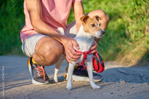 Fotografie Man crouches beside a small terrier and holds its harness on an outdoor path