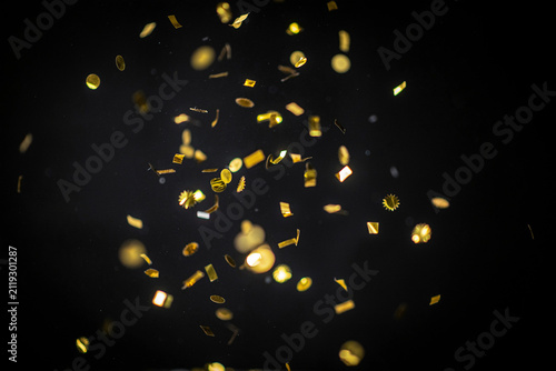 Shimmering golden glitter confetti pieces in various shapes including circles squares and starbursts falling through the dark air creating a festive and celebratory atmosphere of joy