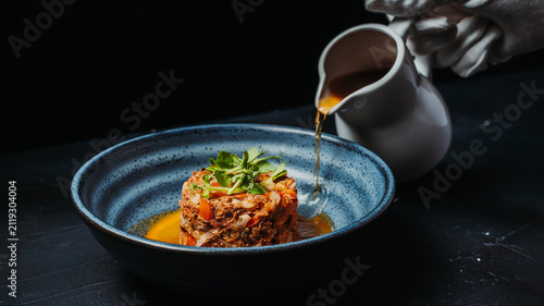 Chef pours rich sauce over a gourmet steak tartare served in a ceramic bowl with fresh microgreens. Elegant fine dining presentation against a black background.