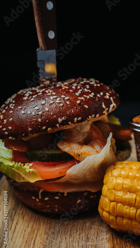 Gourmet shrimp burger served with crispy French fries, grilled corn on the cob, and dipping sauce on a wooden serving board against a black background.