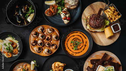 Overhead view of assorted gourmet dishes including appetizers, soups, burgers, seafood, and desserts served on a dark restaurant table.