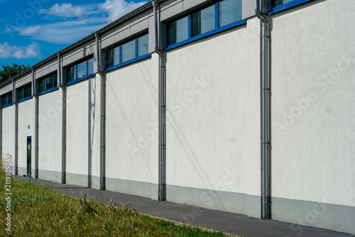 Long facade of an industrial warehouse with metal downspouts and blue upper windows, paved walkway, grass in the foreground, clear blue sky, outdoor shot.