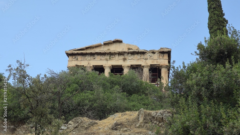 Fototapeta premium Athens, Greece - 8 July 2026. Temple of Hephaestus in the Ancient Agora seen above olive trees and shrubs, with Doric architectural columns, interior scaffolding, a cypress tree, and clear sky.