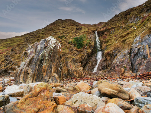 Small waterfall flows down mossy cliffside. Pebble beach stretches beneath rocky outcrop. Wildlife-free nature scene with greenery and water. No animals or plants identified in image. Nature beauty.