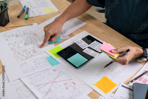 UX UI designer working on wireframes for a website and mobile app prototype, surrounded by sketches of user-flow and design tools, in concept of website and mobile application design concept.