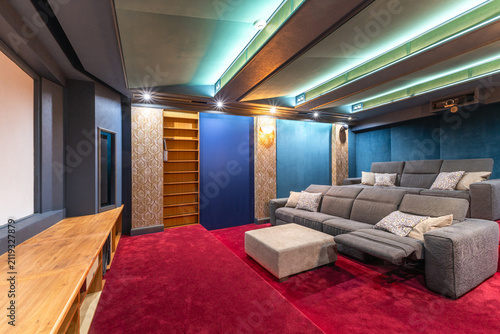 A home theater features gray seating arranged for comfort and viewing. The space includes a large screen, decorative walls, and soft lighting. Shelves are visible in the background