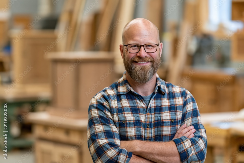 Obraz premium Smiling Bearded Craftsman with Glasses in Wood Workshop Surrounded by Furniture Pieces