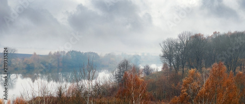 Overcast autumn lake surrounded by trees under foggy sky with heavy clouds
