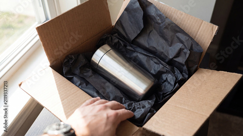 Hands opening a cardboard parcel with an insulated steel bottle beside a window