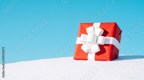 Red wrapped gift box with a white ribbon resting on fresh bright snow