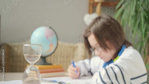 Teenager sitting at desk writing homework in notebook, hourglass time management, education, knowledge and writing, learning process, exam prep, child training, concentration and discipline sand timer