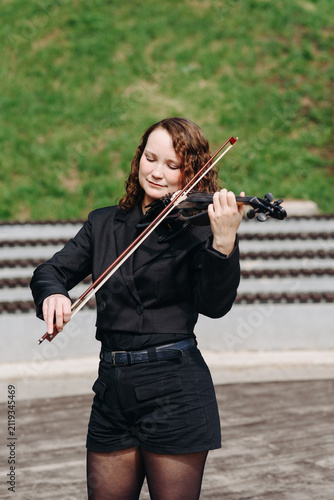 violinist outdoors, young beautiful redhead caucasian woman in jacket playing black violin in city park with green grass and trees in sunny summer day, classic music concept