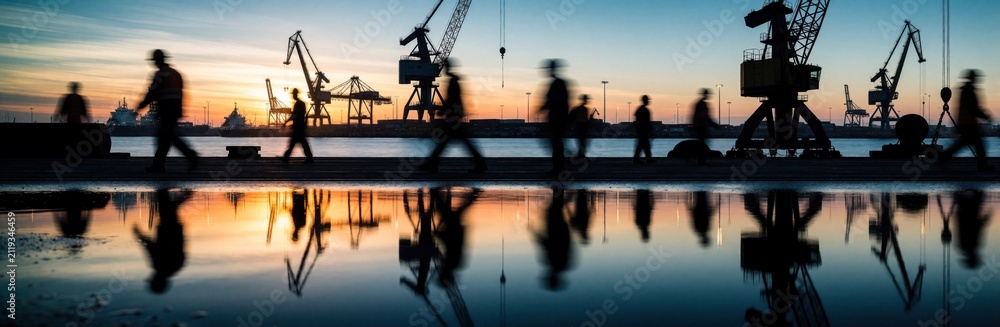 Obraz premium Port workers walk at sunset near large industrial cranes. Laborers move across shipping dock by ocean water reflections. Busy harbor scene during evening shift change features silhouettes of heavy