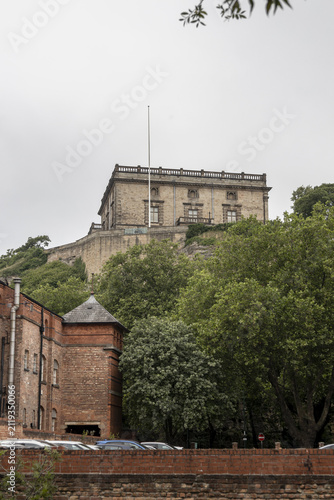 View of the historic Nottingham Castle perched on a rocky hill surrounded by lush green trees and a brick wall in the foreground in Nottingham, England, United Kingdom.
