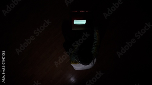 Person sitting in a dark room, facing a glowing retro television broadcasting a vintage screen display, representing media consumption and nostalgia