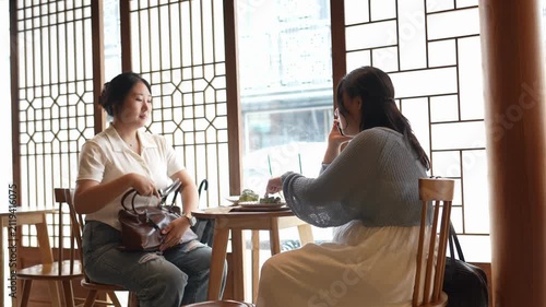 Two women sit at a table, sharing a quiet conversation. Soft light streams through traditional window panels. One woman smiles, the other listens attentively. Serene atmosphere suggests calm