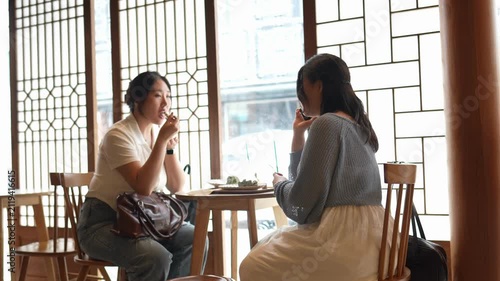 Two women sit at a table, conversing. Natural light streams through lattice windows. Their expressions show calm engagement. The setting evokes quiet intimacy