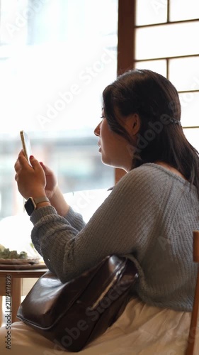 Woman sits by window, sunlight glows softly. She waves gently, then holds phone, seems relaxed. Brown bag rests on her lap, watch glints subtly. Cozy cafe moment, quiet, peaceful, thoughtful