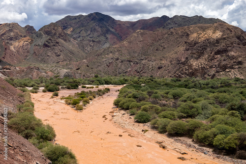 Scenic panoramic view of a wide muddy brown river flowing through a dry valley with green shrub and red mountains on a sunny day in Salta, Argentina, South America. Vivid, saturated colors