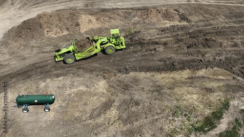 Earth scraper dumps a full load of soil while driving uphill at an active construction site, with heavy equipment moving earth and a burst of black exhaust under engine load.

