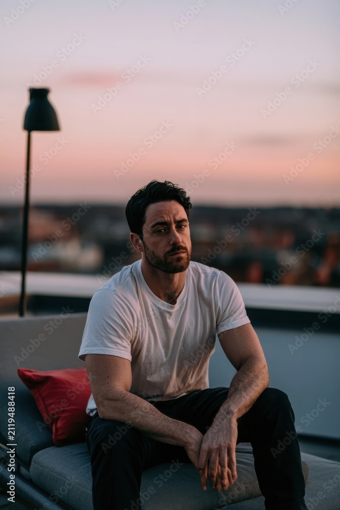 Fototapeta premium Pensive man in white t-shirt sitting on rooftop at sunset