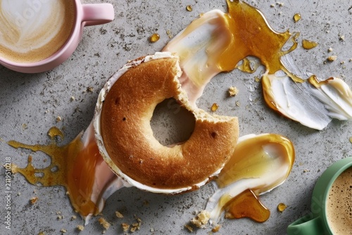 High-angle view of a half-eaten glazed donut on a grey concrete surface with spilled caramel sauce and two ceramic coffee cups