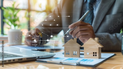 Real estate agent wearing a professional suit holds a pen while analyzing digital housing market data alongside wooden house models to facilitate property investment discussions in an office.