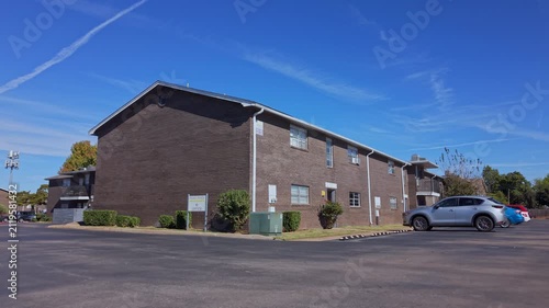 Sunny daytime view of a brick multifamily housing complex with parked cars, shrubs, and clear signage.