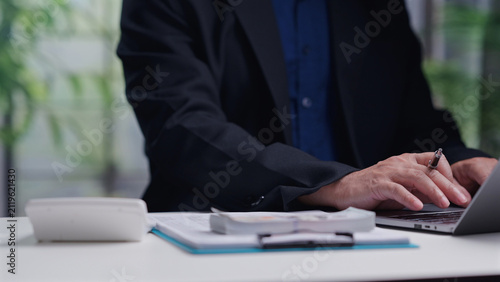 Male accountant wearing dark suit operates laptop keyboard, managing corporate financial budget, calculating taxes, counting cash money stack, planning business investment inside office.