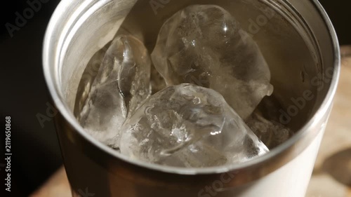 Close-up top angle view into stainless steel tumbler cup with melting ice cubes in golden warm light on wooden table in cafe