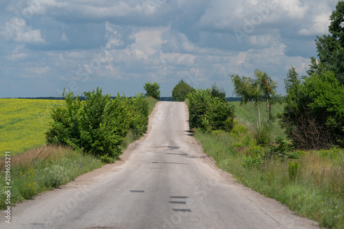 Rural country road rising over gentle hill between green fields and yellow wildflowers beneath dramatic cloudy sky, creating quiet open landscape with strong depth and inviting travel mood