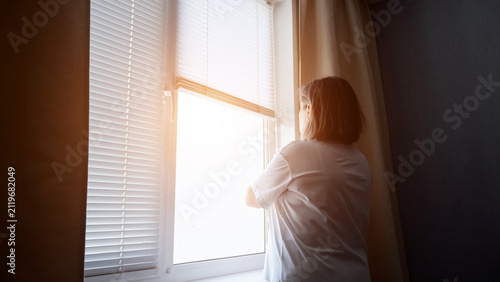 Woman standing by a window with blinds, looking at bright sunlight from outside, contemplating thoughts of future and opportunities with a sense of hope and new beginnings