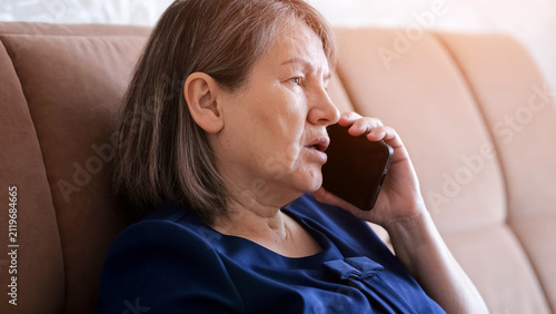Senior woman at home sitting on a sofa, holding a mobile phone to her ear and talking with a concerned expression, communicating important news or receiving advice