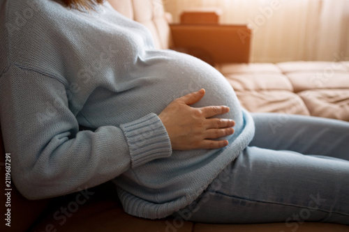 Expectant mother in soft home clothes lies on a couch, gently holding her belly. Warm natural light and a calm, comfortable living room create a peaceful maternity mood