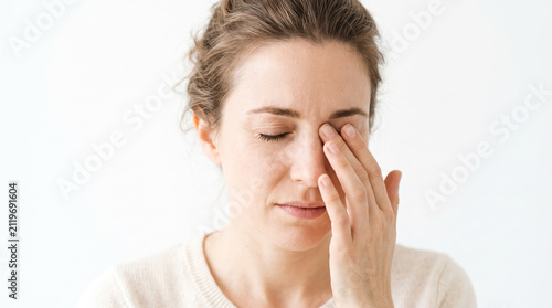 a woman touching her eye gently, blinking in irritation, clear focus on expression and hand gesture, minimalistic white backdrop