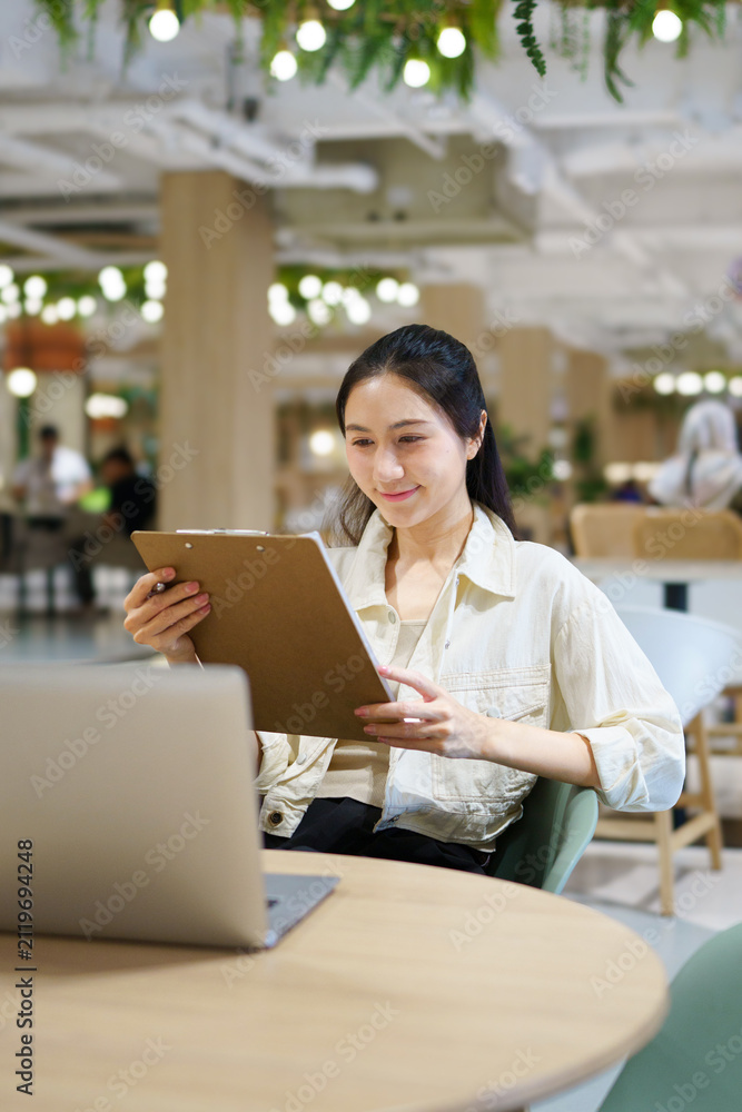 Fototapeta premium Young Asian Woman Reviewing Documents on Clipboard at Modern Co working Space with Laptop