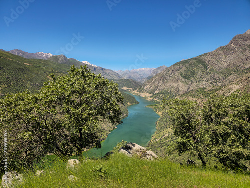 Verdant spring valley enfolds a crystalline turquoise river beneath towering forested hillsides, snow-dusted peaks crown the distant horizon, serene wilderness glowing with fresh mountain light.
