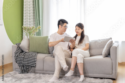 Fotografie A young Asian couple sits on a sofa in a bright living room, jointly viewing a tablet and interacting casually