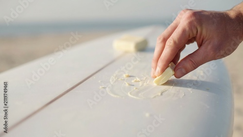 Hand applying sunscreen on a white surfboard at the beach with ocean in background