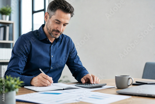 A focused man reviews documents, writes notes and uses a calculator while creating a personal financial plan at his modern home office desk.