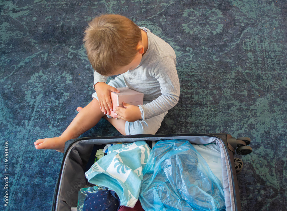 Naklejka premium Cute little boy helping to pack a large black suitcase indoors before a family trip. Toddler playing near open luggage filled with clothes and travel essentials. Highlighting the concept of family .