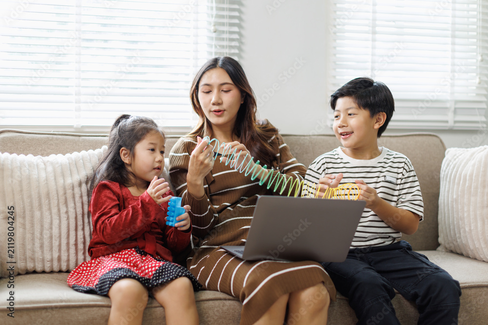 Fototapeta premium Asian mother engaging with her two young children at home. All sitting comfortably on a sofa. Utilizing a laptop while interacting with a plastic spring toy