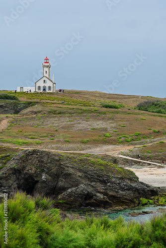 A white lighthouse with a red lantern stands atop grassy, rocky cliffs overlooking a calm coastal cove. Visitors walk the trails while the muted sky and green scrub create a peaceful, remote mood.