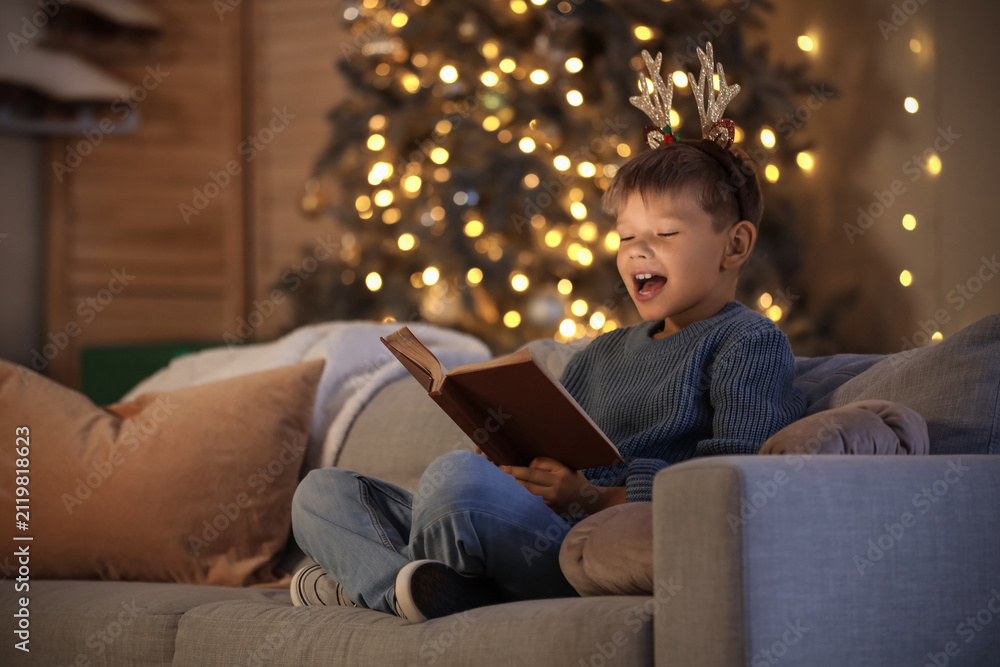 Fototapeta premium Cute little boy reading Christmas story on sofa at home in evening