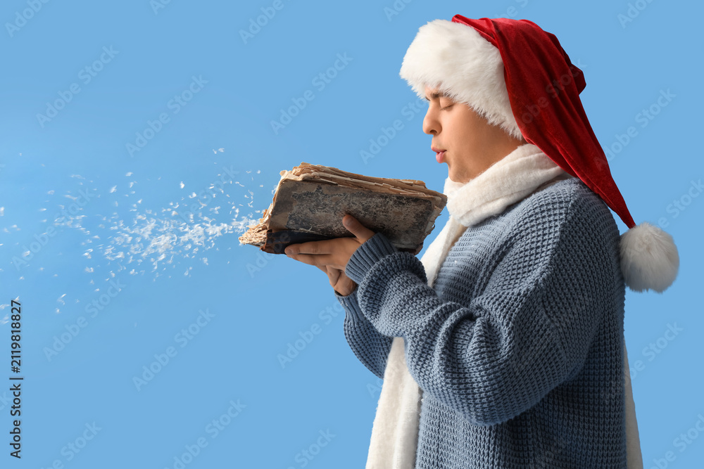 Fototapeta premium Young man in Santa hat and with book blowing snow on blue background