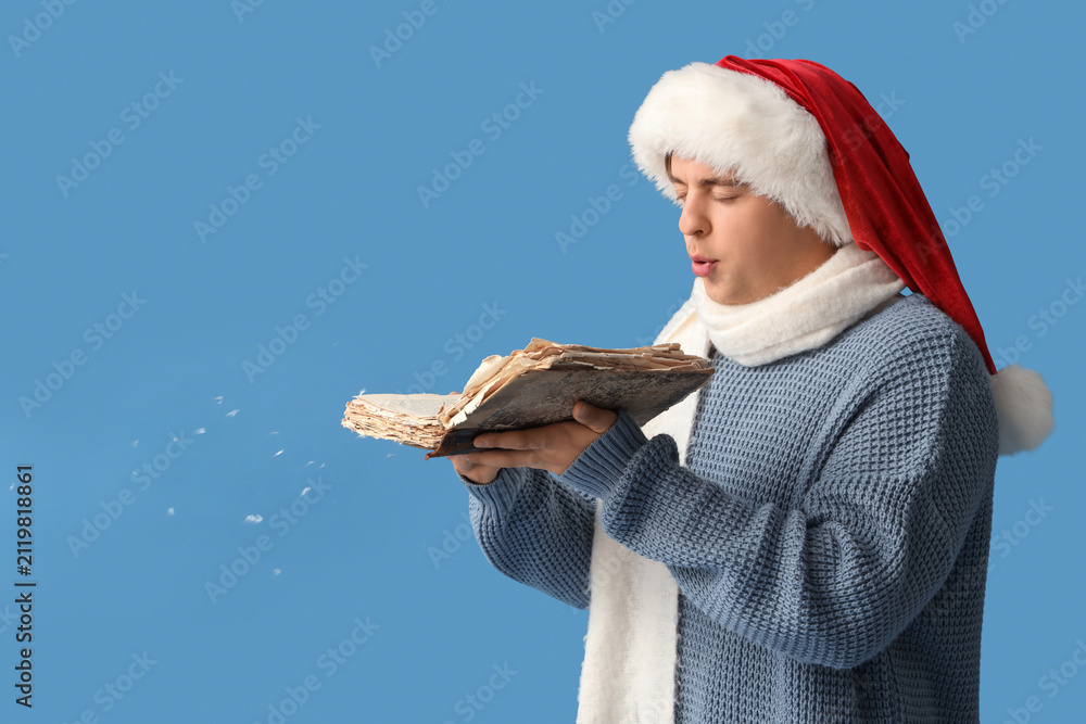 Fototapeta premium Young man in Santa hat and with book blowing snow on blue background