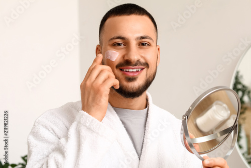 Foto Young man with mirror applying cream on face in bathroom, closeup