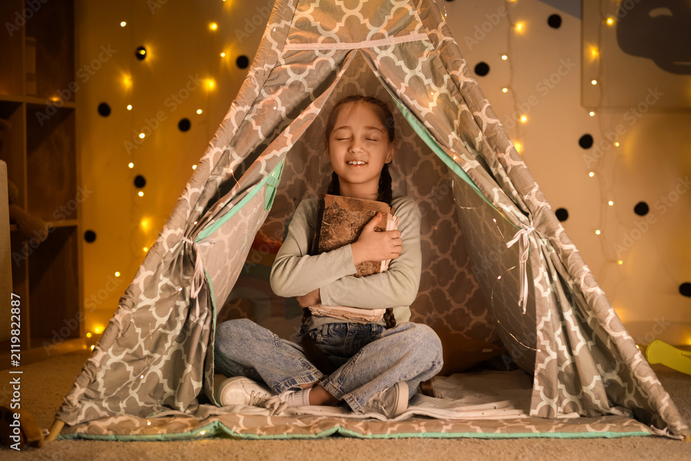 Fototapeta premium Cute girl with adventure book sitting in play tent at night