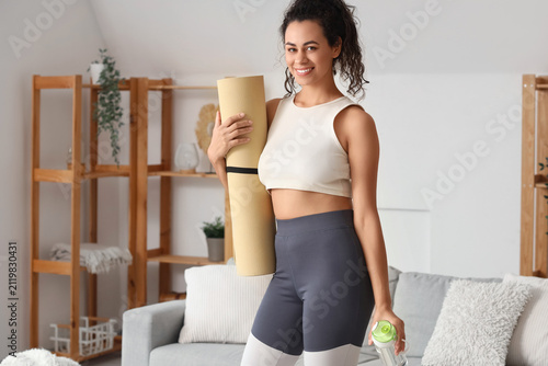 Sporty young African-American woman with fitness mat and water bottle at home